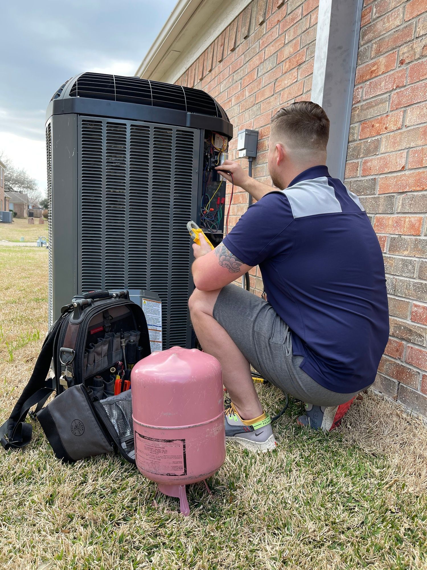 A man is kneeling down in the grass working on an air conditioner.