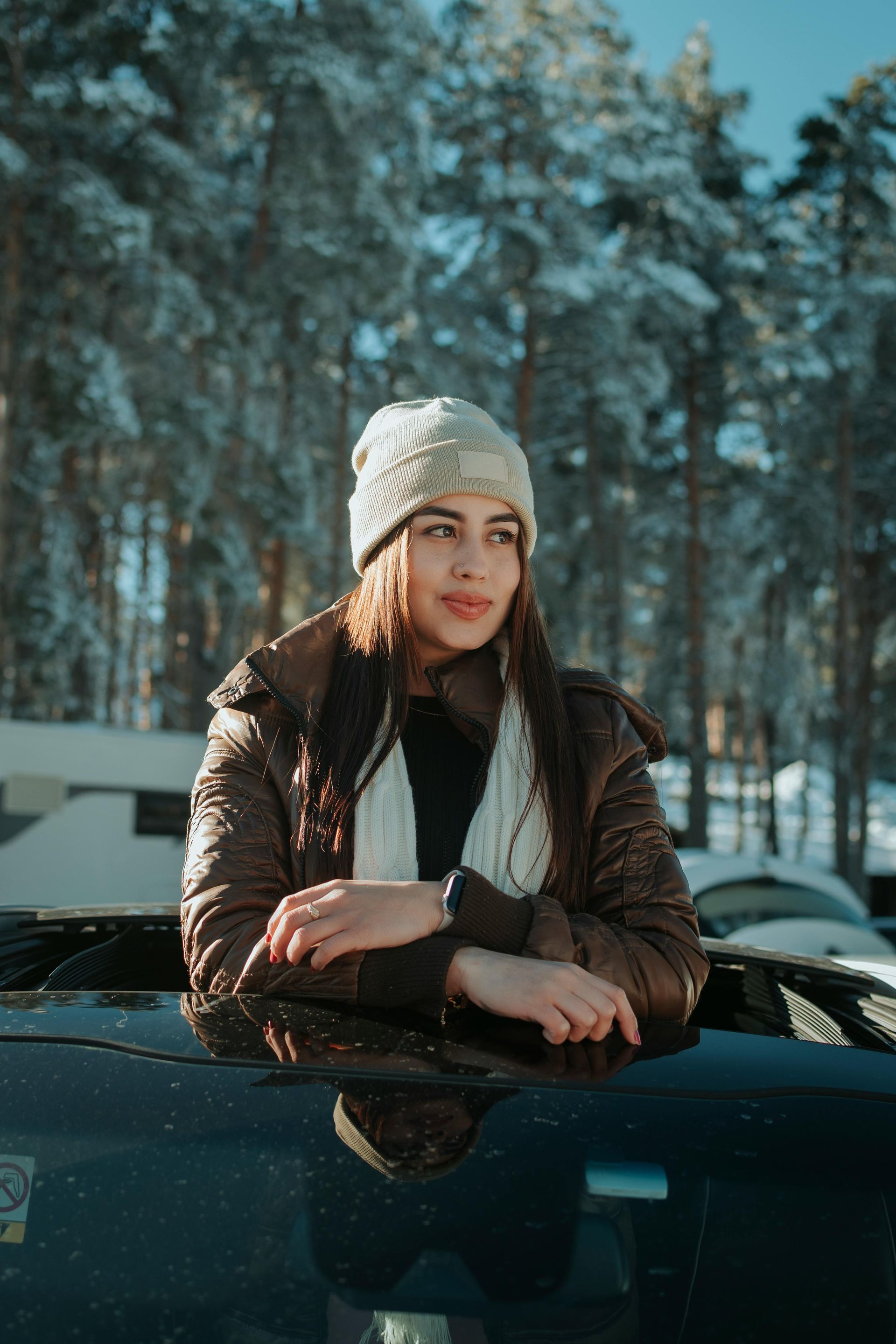 Woman in a winter hat and brown jacket leans on a car, looking to the side in a snowy forest.