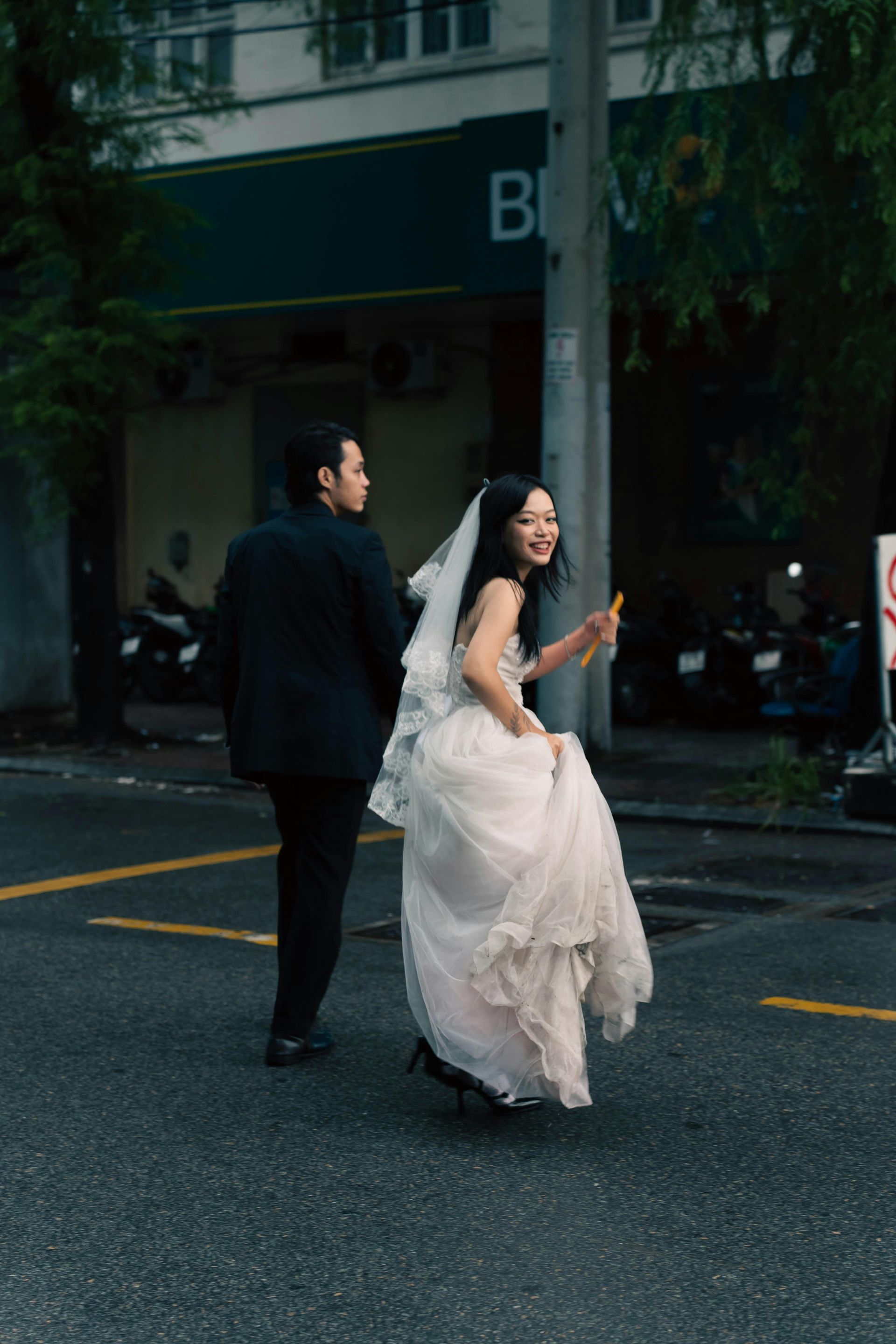 Bride and groom crossing a street, bride smiling and holding a bouquet, groom walking.