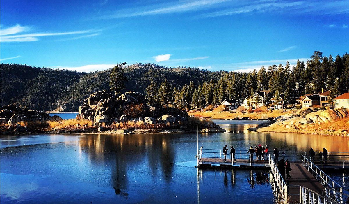 Lake view with a wooden dock, people, rocks, trees, and homes under a blue sky.