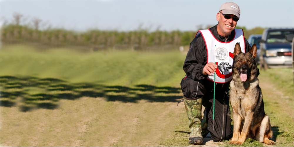 A man kneels with a German Shepherd. Both are on a grassy field. The dog is panting, tongue out.