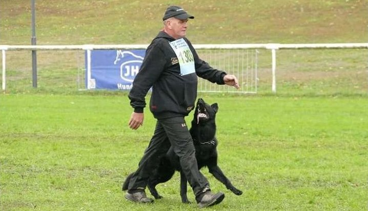 Man in black clothing walking with a black dog on a grassy field; they appear to be in a competition.