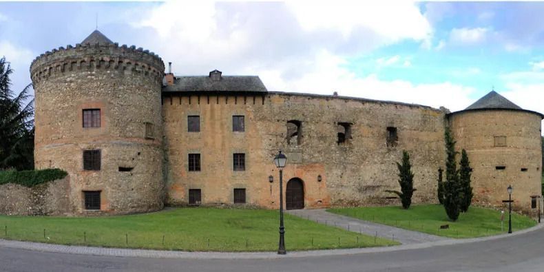 Stone castle with cylindrical towers and arched entrance. Green lawn, trees, and cloudy sky.