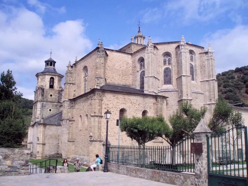 Stone church building under a partly cloudy sky. People sit on a bench near a wrought-iron fence.