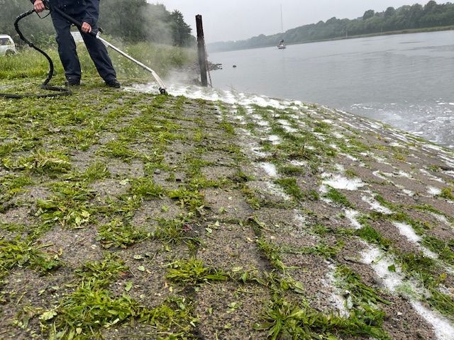 Ein Mann bespritzt mit einem Schlauch Gras auf dem Boden in der Nähe eines Gewässers.