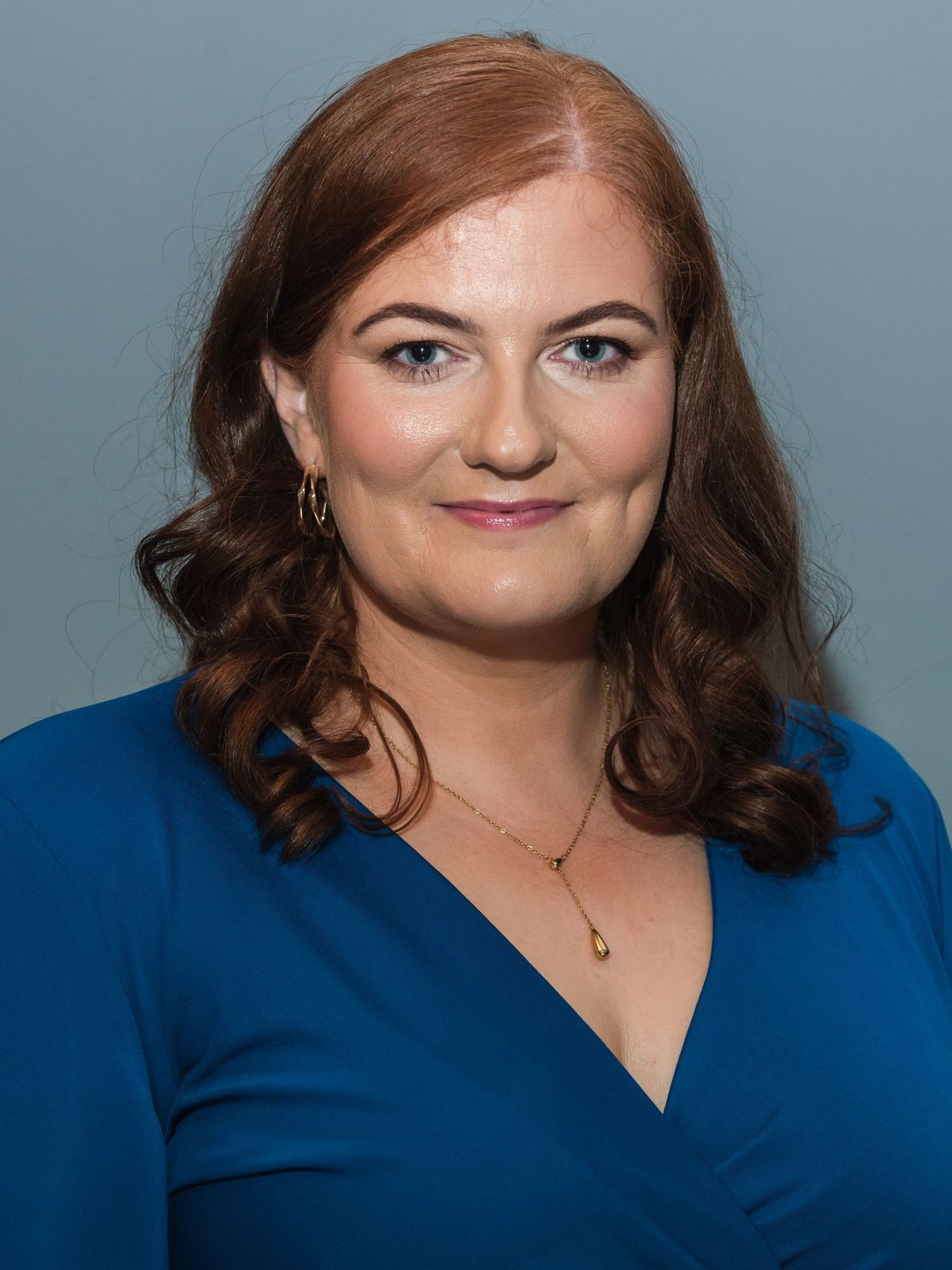 A woman with red hair is wearing a blue shirt and a gold necklace.