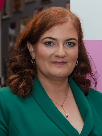 A woman with red hair is wearing a green jacket and a gold necklace.