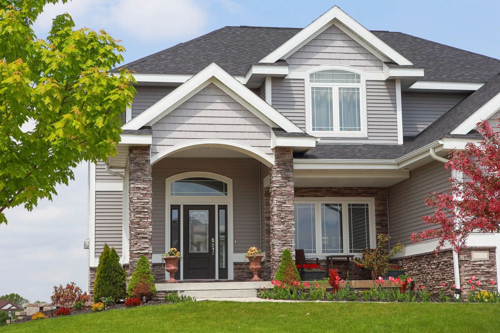 Two-story gray house with stone columns, arched porch, green lawn, and colorful landscaping.