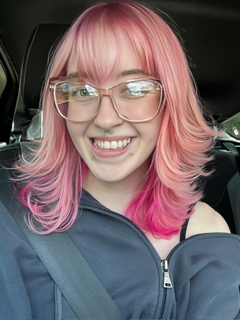 A woman with pink hair and glasses is smiling in a car.