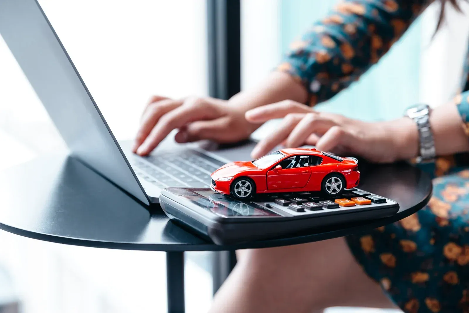 Person using laptop with red toy car on calculator on a table.