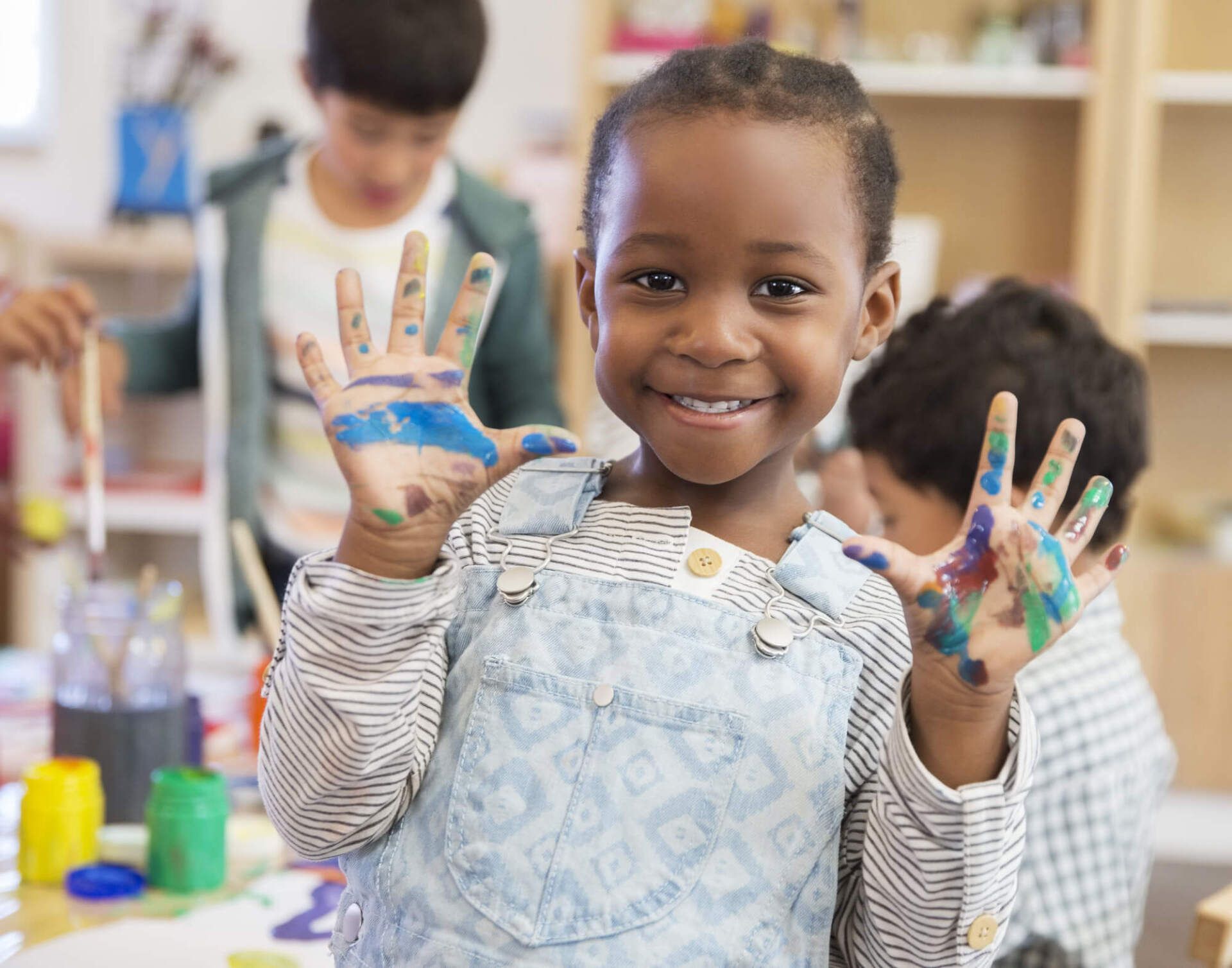 A little girl smiling and holding up her paint covered hands to show the camera