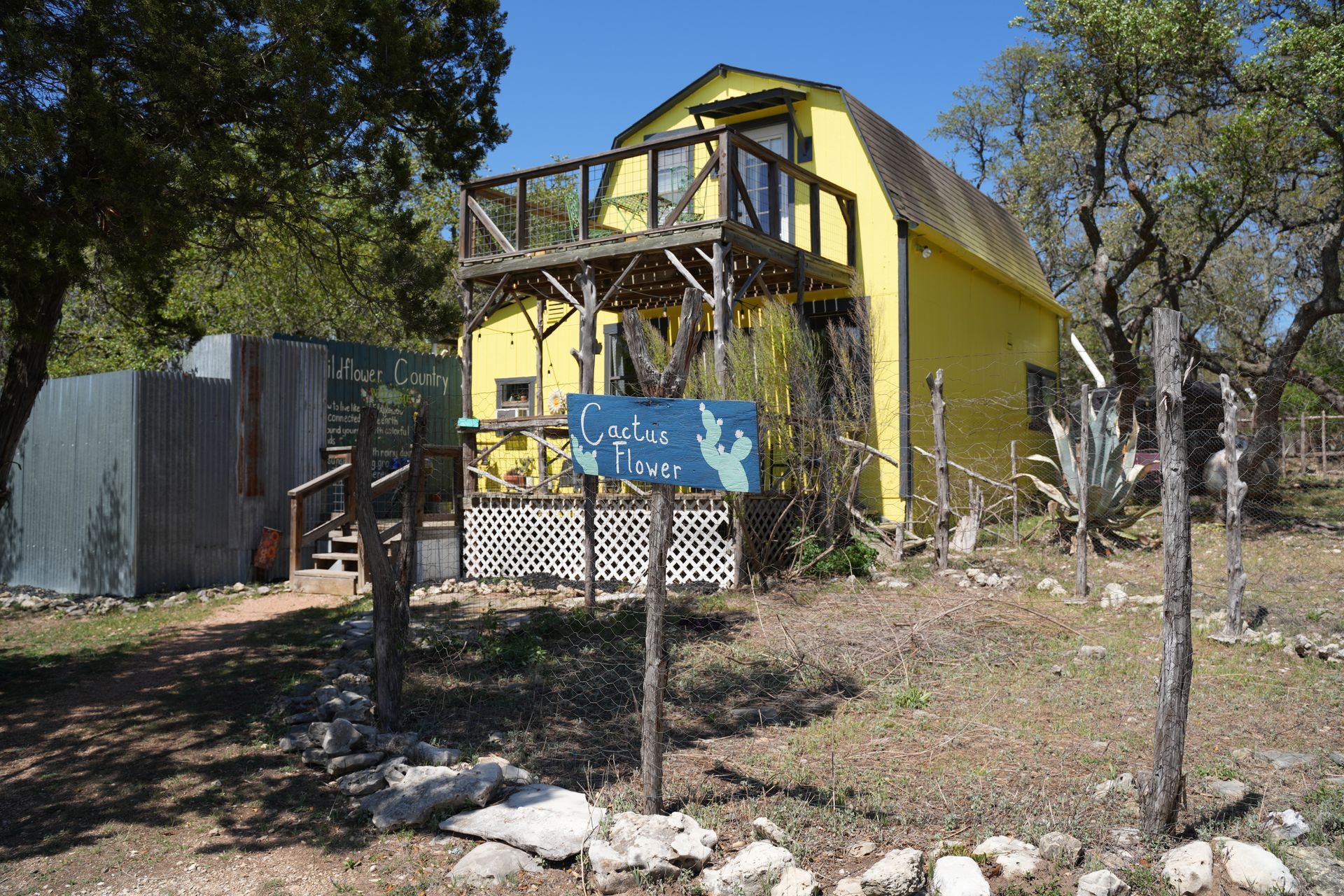 A bright yellow barn-style house with an upper wooden deck sits in a clearing surrounded by trees and a rustic fence.