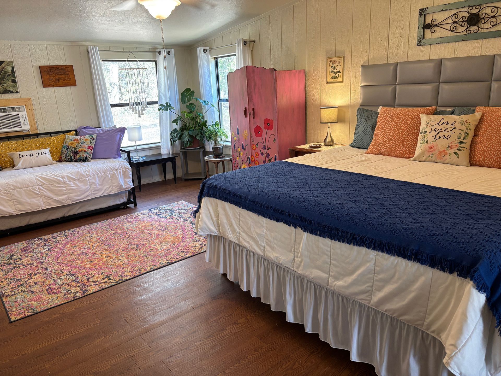 A bedroom with a king-sized bed, a daybed, a pink folding screen, and a patterned area rug on a wooden floor.