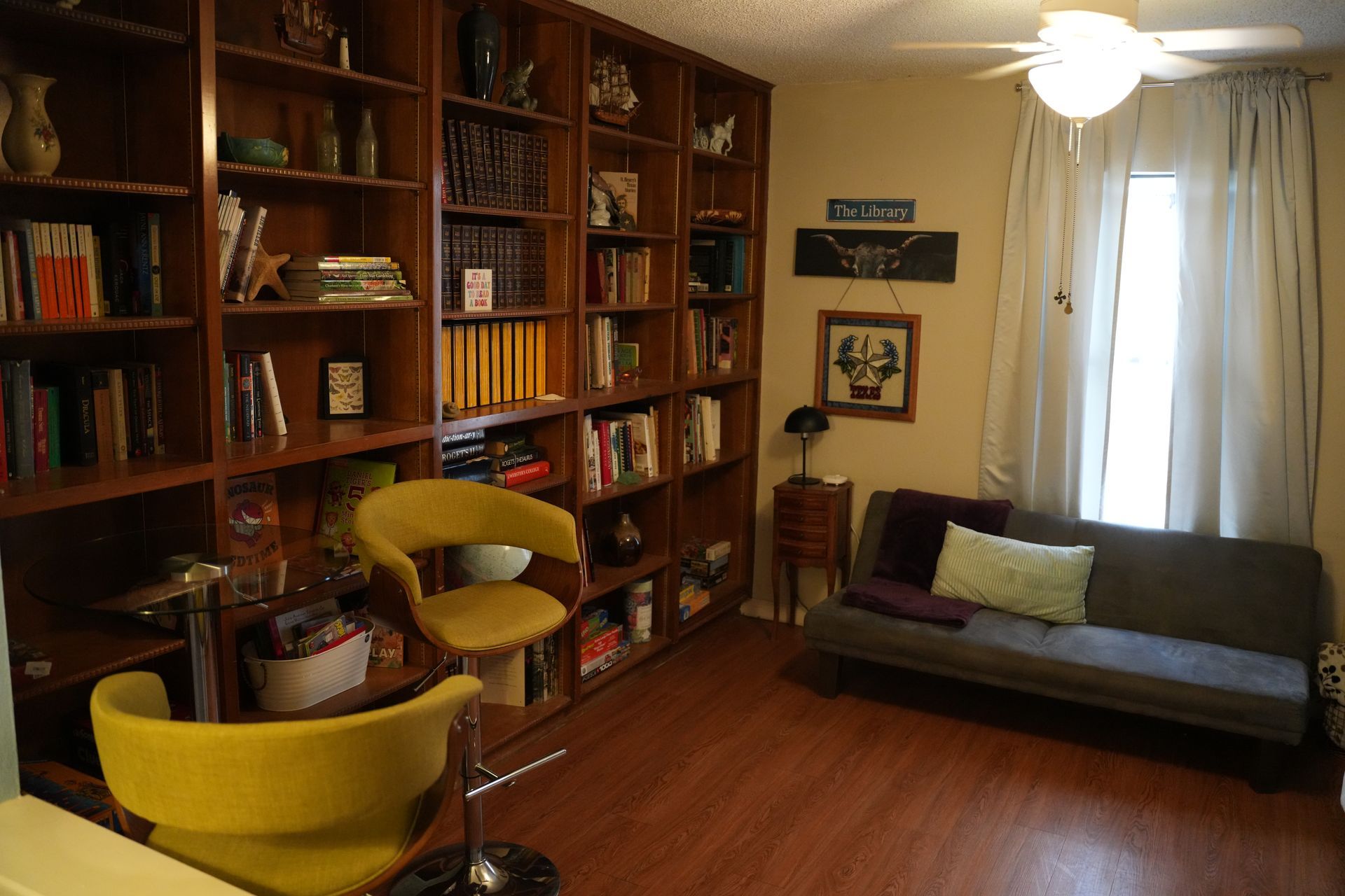 A home library with a large wooden bookshelf, two yellow swivel chairs, a gray sofa, and a window with white curtains.
