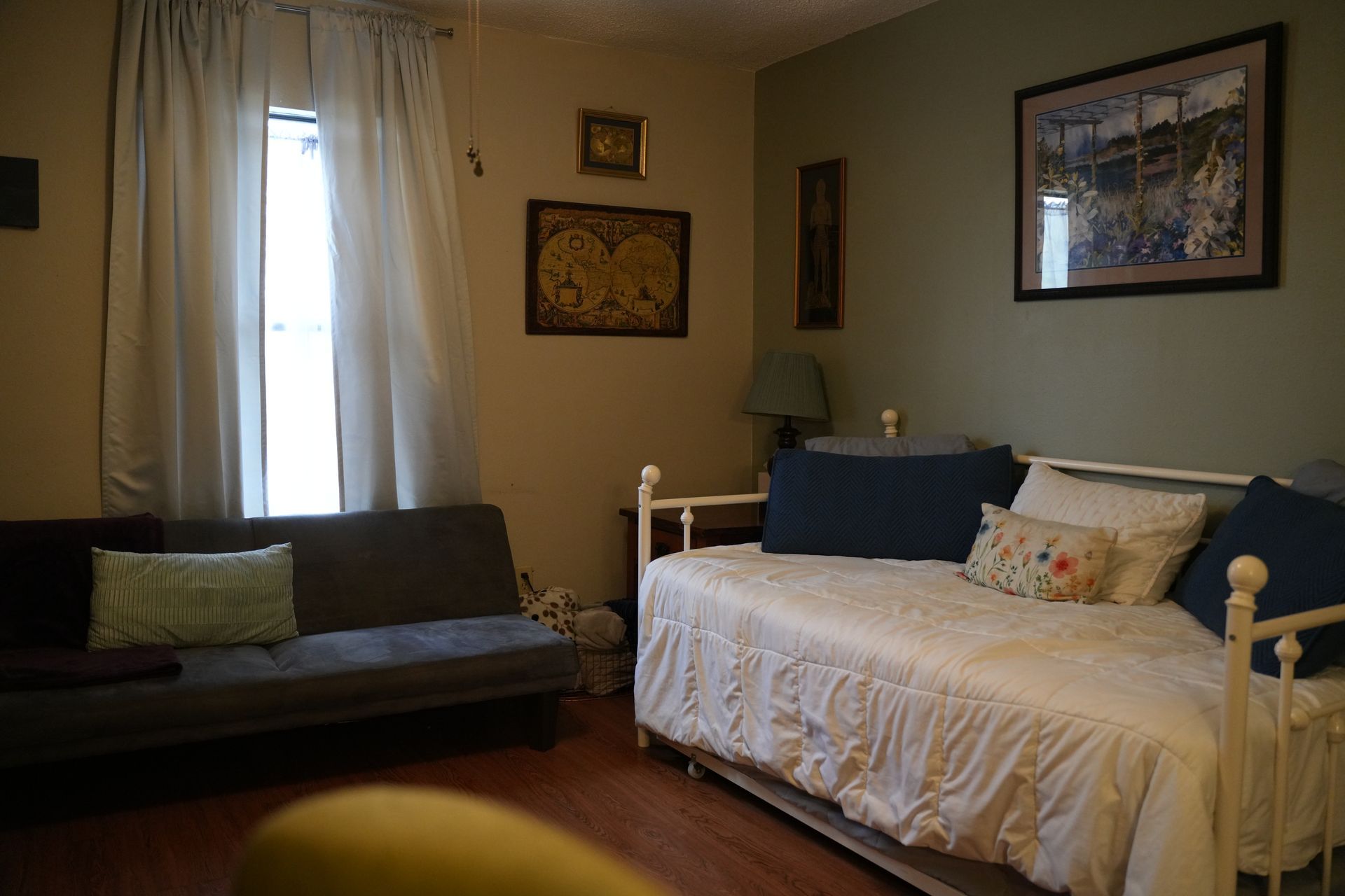 A cozy room featuring a daybed with blue and floral pillows, a gray futon, window curtains, and wall art.
