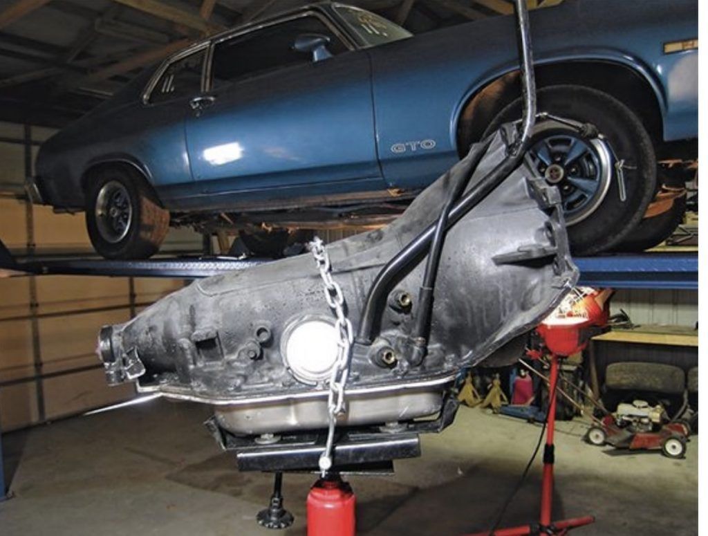 A car transmission being lowered from a blue muscle car on a lift in a garage | Loyola Service