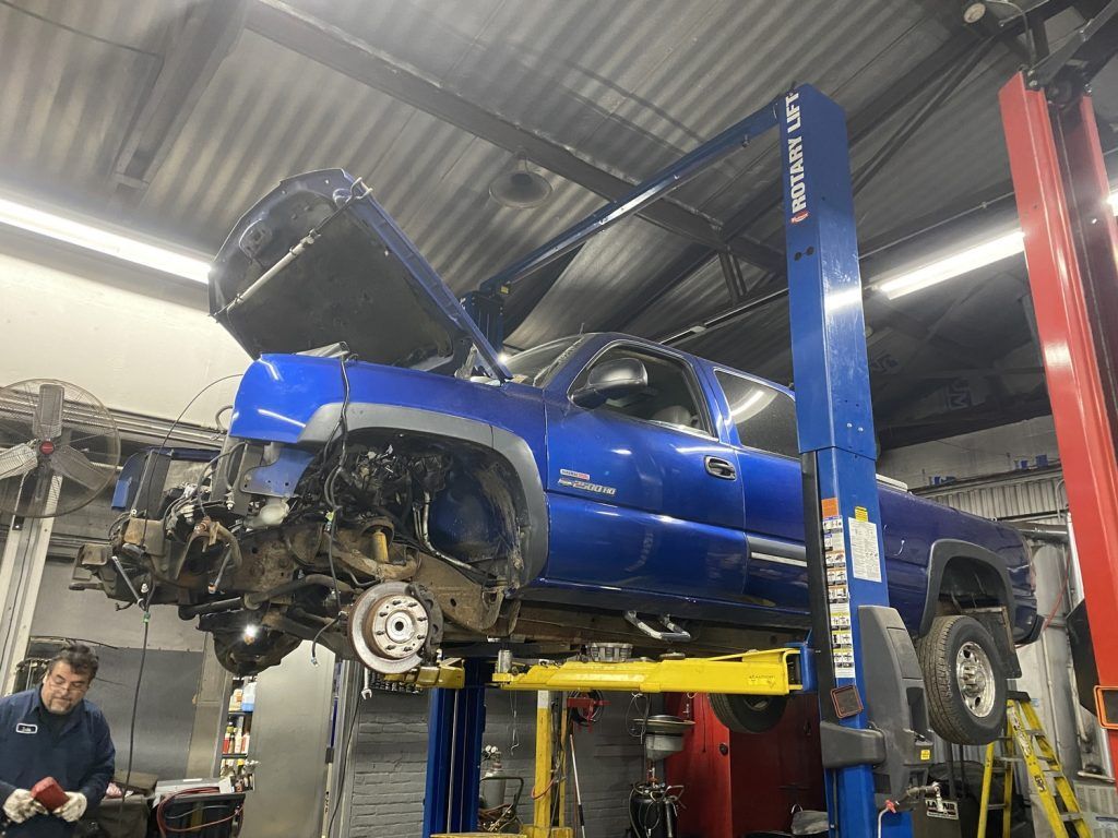 Blue truck on a lift in a repair shop, hood open, front end disassembled, mechanic in the foreground. | Loyola Service