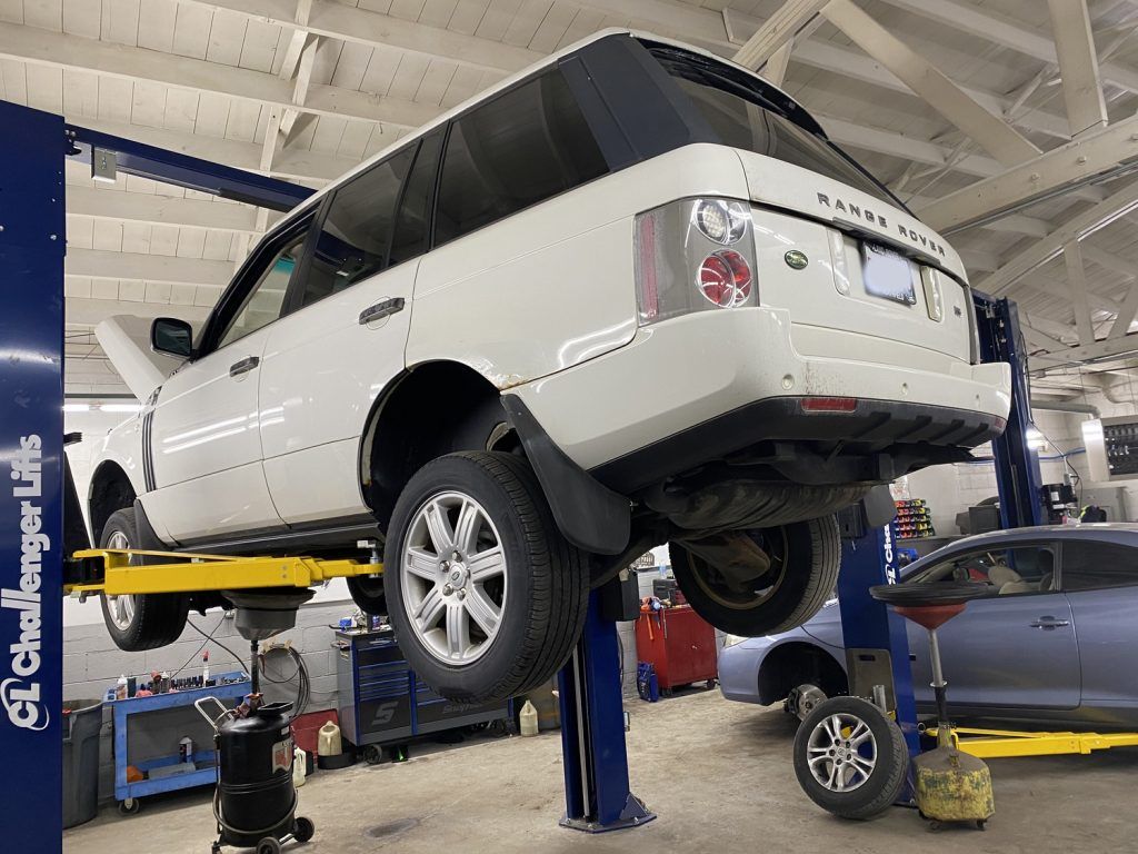 White SUV on a lift in a garage, wheels off the ground.  Tools and a car are visible nearby. | Loyola Service