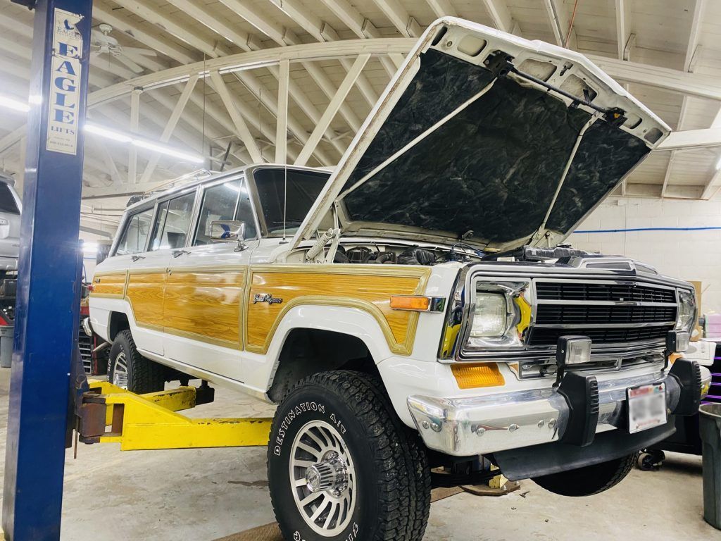 A classic white and wood-paneled Jeep Wagoneer with the hood open, on a lift in a garage. | Loyola Service