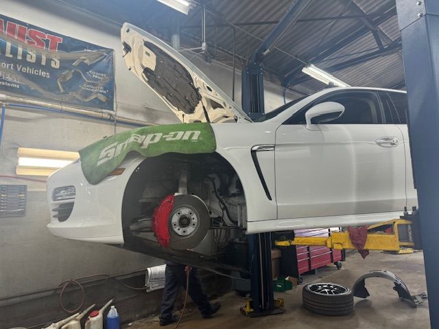 White car on a lift, hood open, front tire removed, red brake rotor visible. In a repair shop. | Loyola Service