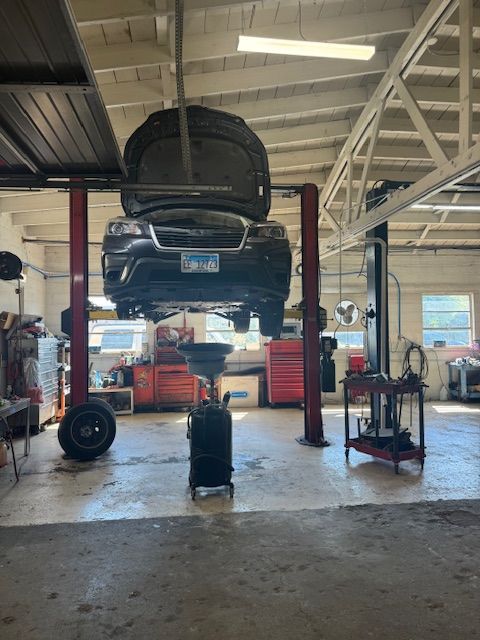 Car on a lift in a garage. Hood open, oil drain pan below. Red tools and equipment visible. | Loyola Service