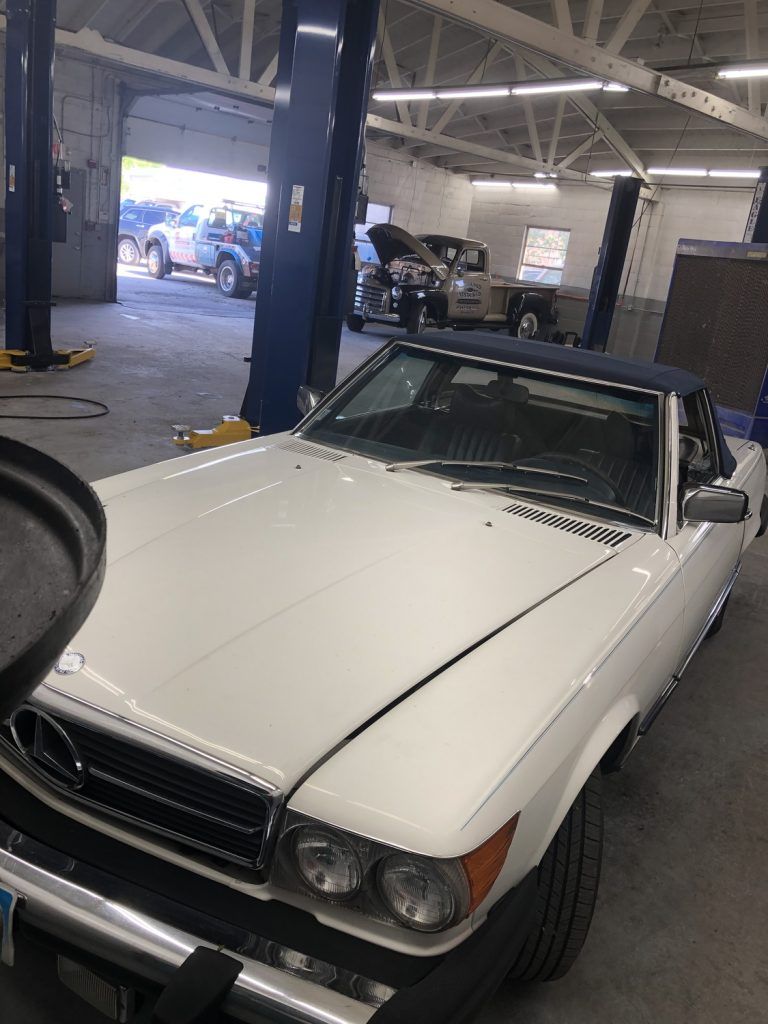 White Mercedes convertible in a mechanic's shop, blue lift arms visible. | Loyola Service