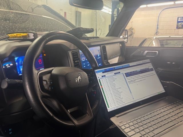 Laptop connected to a Ford Bronco's dashboard for diagnostics in a garage. Steering wheel and gauges visible. | Loyola Service