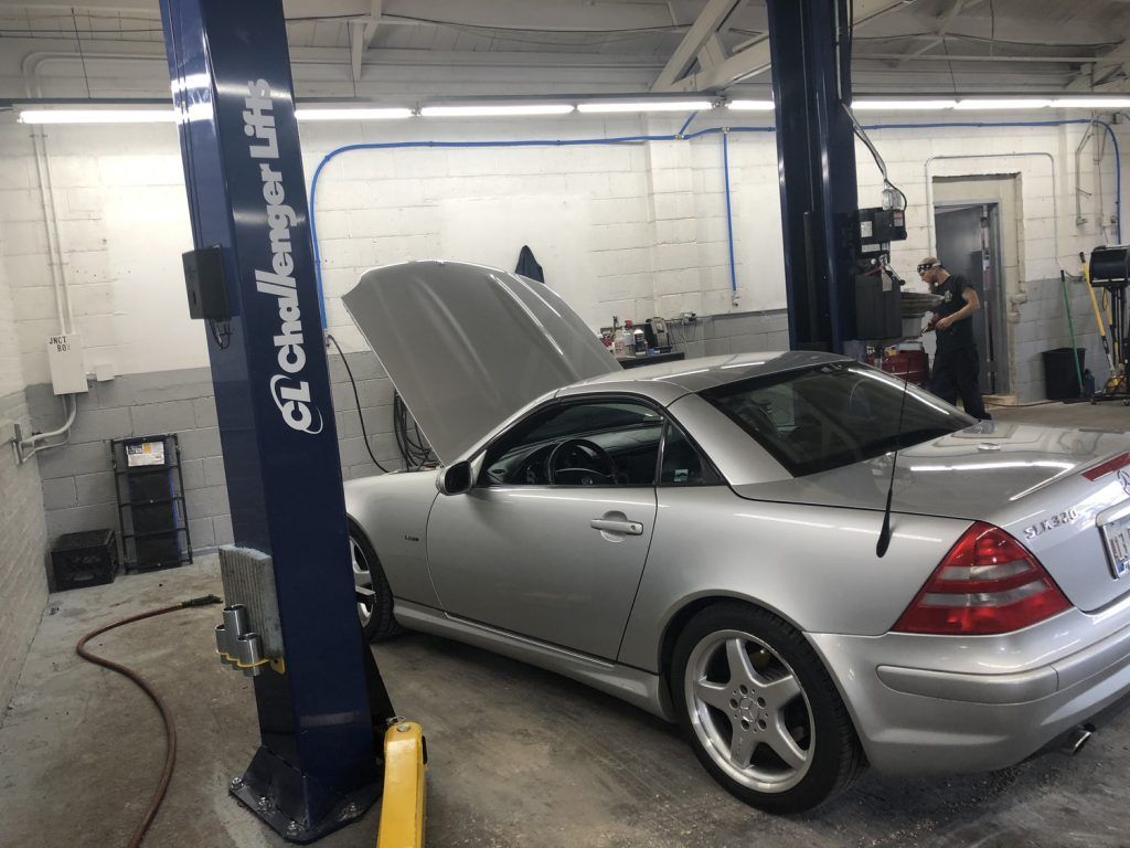 Silver Mercedes sports car on a lift in a repair shop with the hood open, a mechanic in the background. | Loyola Service
