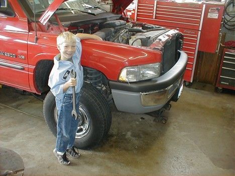 Boy holding wrench stands by the open hood of a red truck in a garage. | Loyola Service