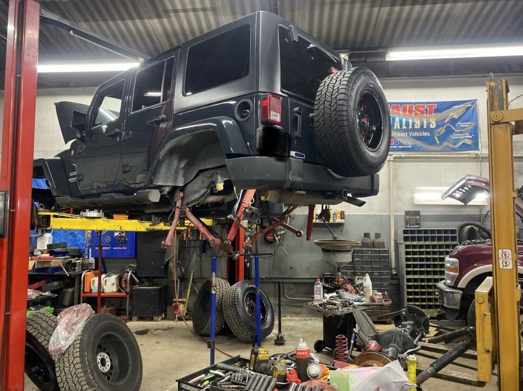 Black Jeep lifted in a repair shop, suspension work underway. Tools and parts scattered around | Loyola Service