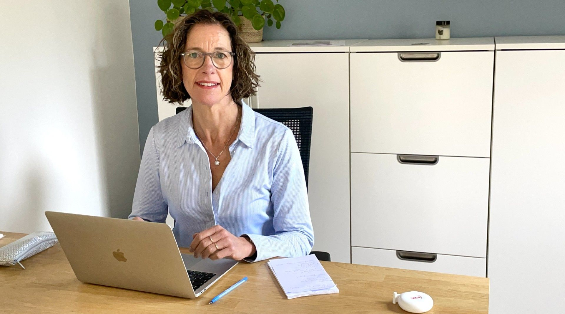 Vrouw in blauwe blouse zittend aan bureau achter laptop. Witte kasten op achtergrond