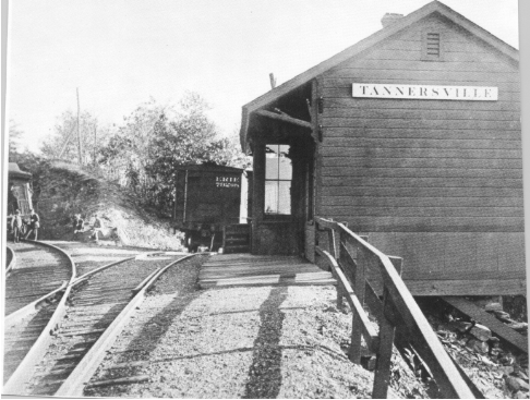 Tannersville train station with tracks, bridge and train car. Building sign above door. Black and white photo.