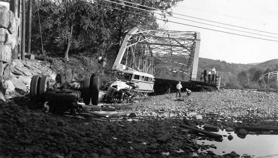 Overturned bus crushed by a collapsed bridge. Debris, rocks, and people surround the wreckage on a gravelly riverbank.