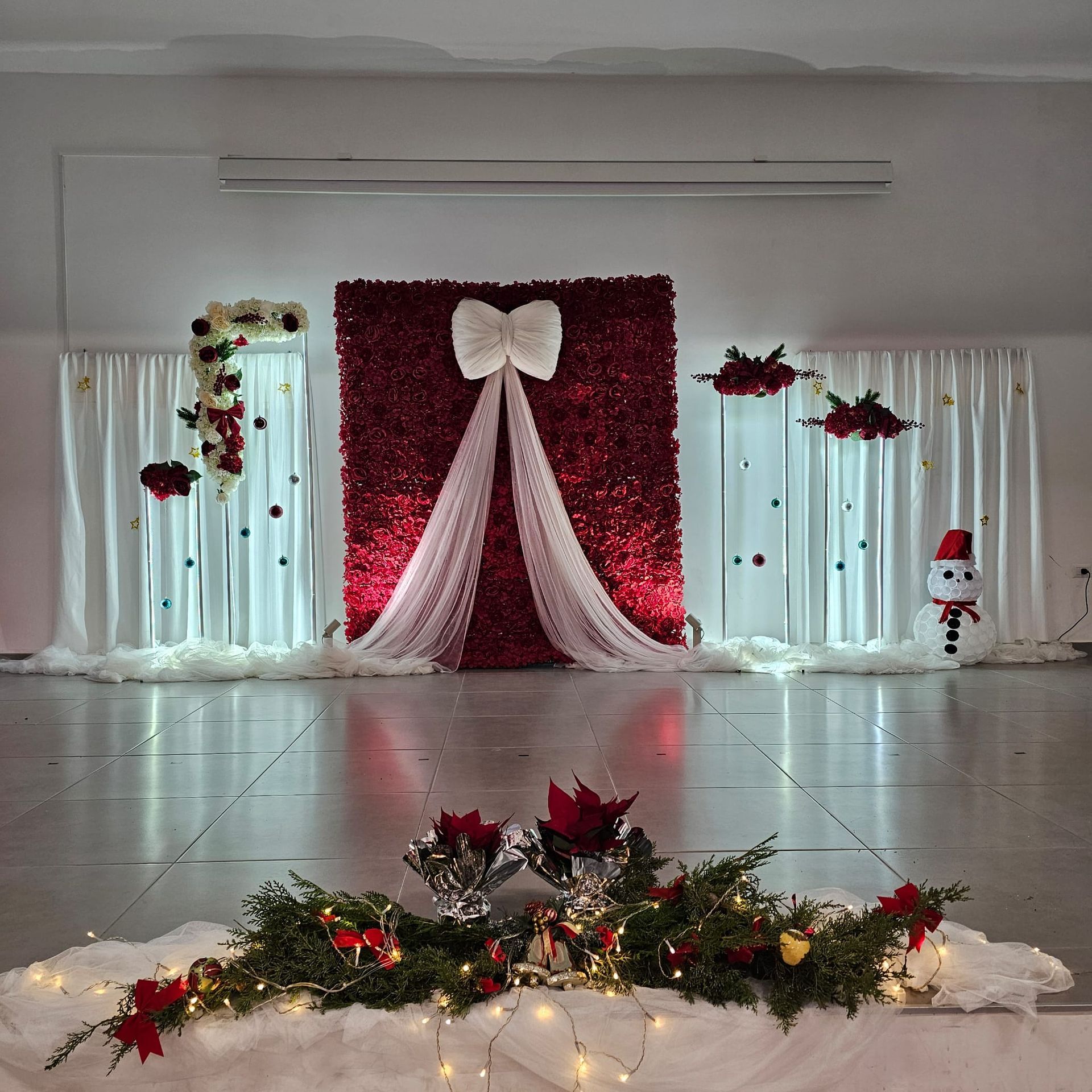 Palco a tema natalizio con parete floreale rossa, fiocco bianco, tende e decorazioni.