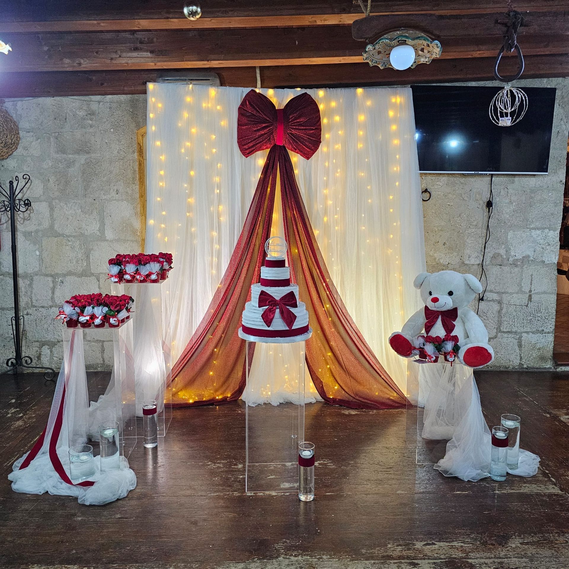Decorazioni nuziali con fiocchi rossi, tessuto drappeggiato, una torta e un orsacchiotto di peluche, il tutto su uno sfondo di luci.