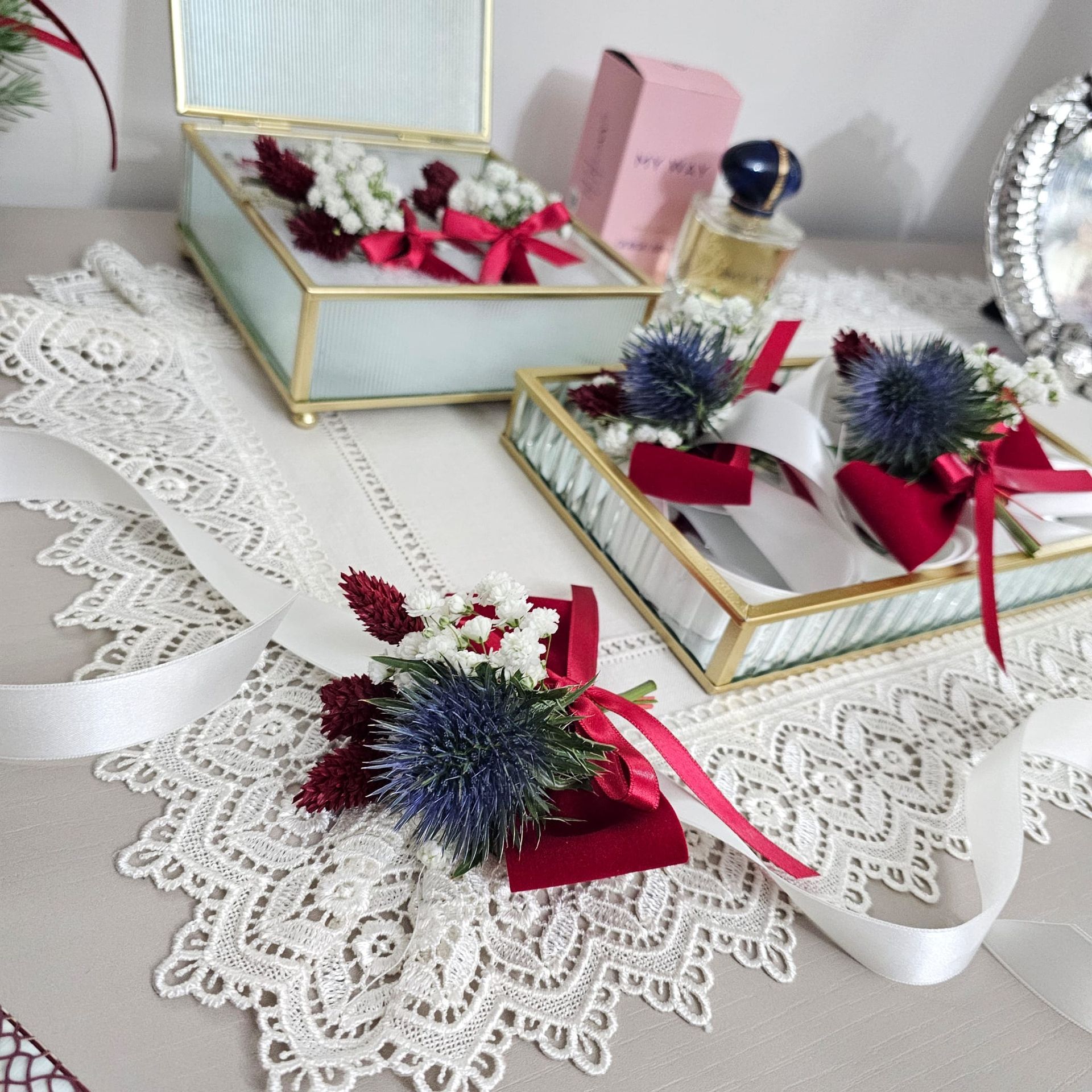 Primo piano di scatole decorative per anelli e composizioni floreali su un centrino di pizzo, con profumo e uno specchio sullo sfondo.