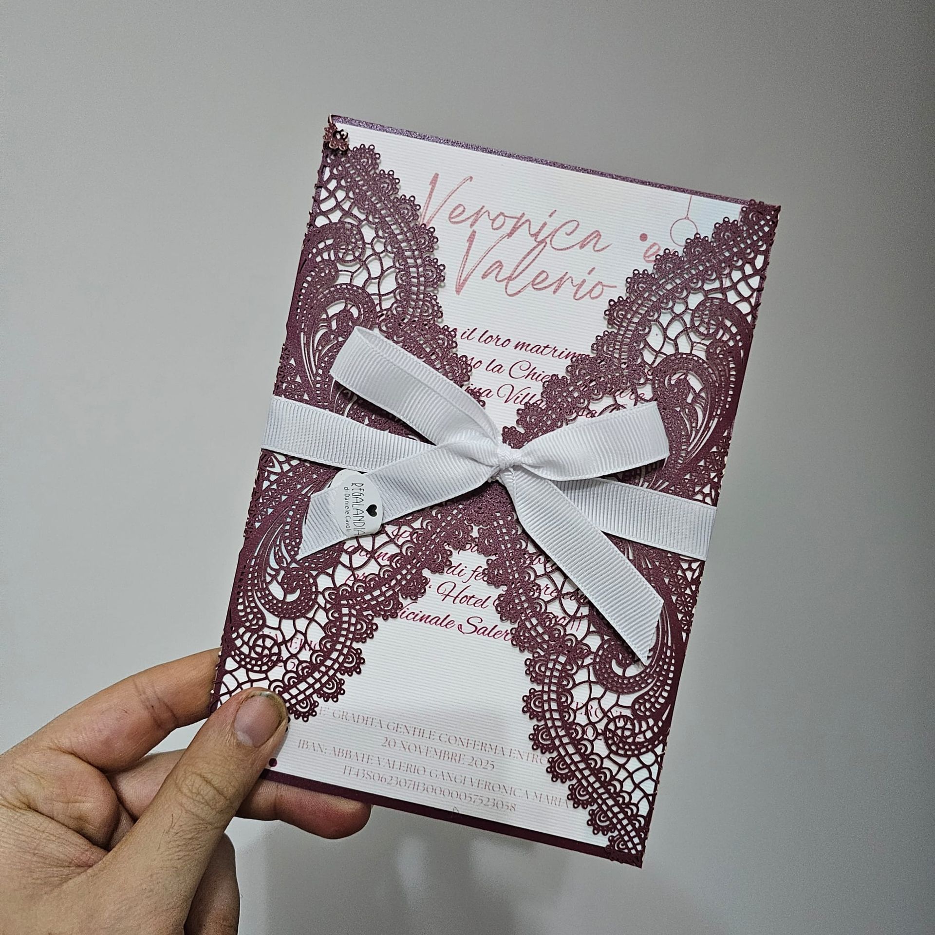 Mano che regge un invito di nozze tagliato al laser color prugna con un nastro bianco.