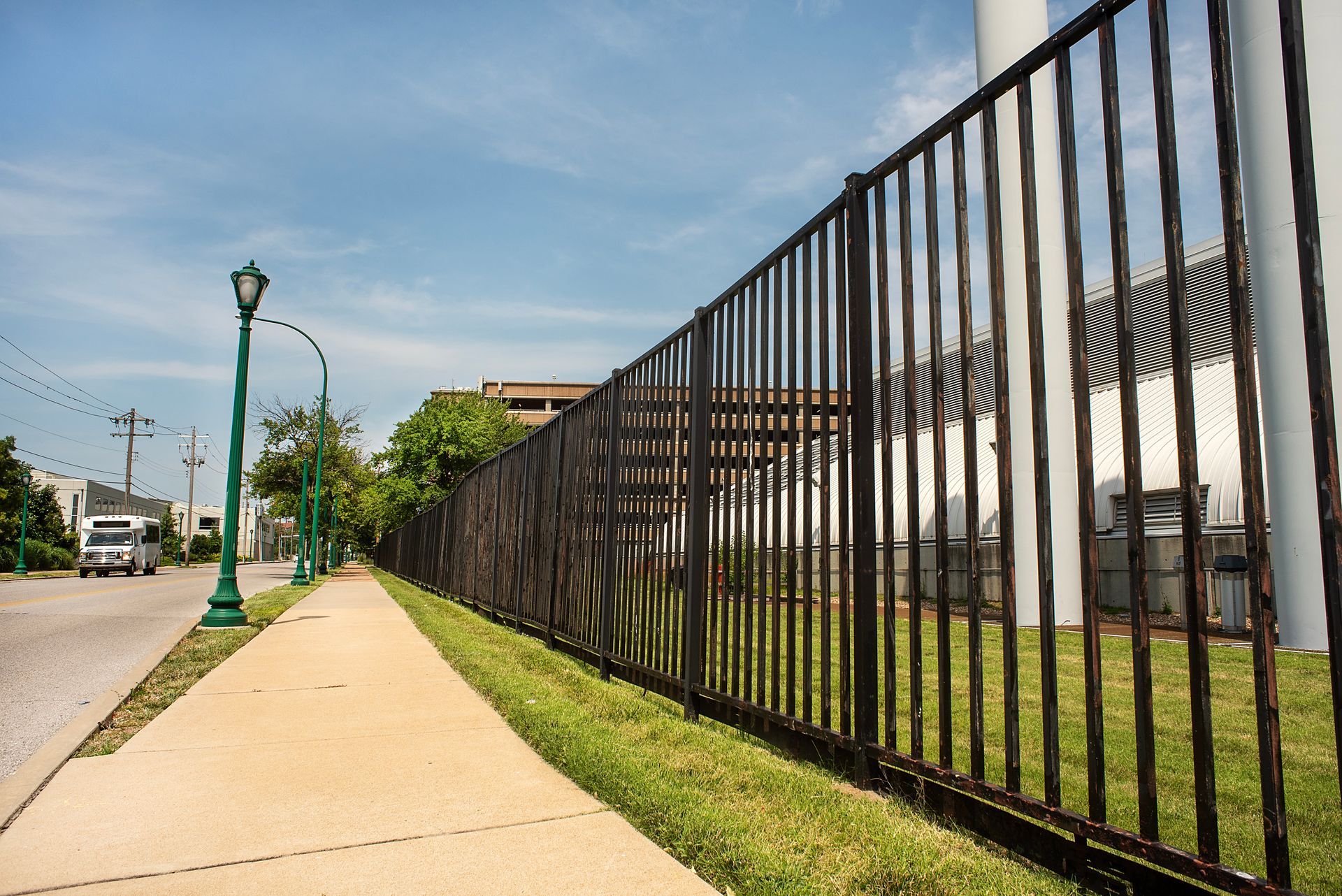 Sidewalk alongside a black fence, a street with a white truck, and a light post under a blue sky.