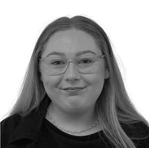 Woman with glasses smiles, wearing a black jacket and shirt, in a grayscale portrait.