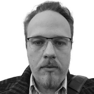 Man with glasses and a goatee, wearing a plaid jacket, looks at the camera.