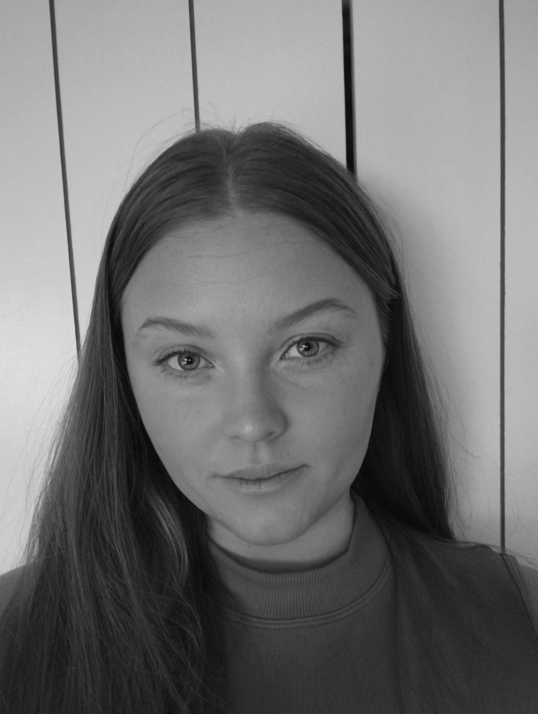Black-and-white portrait of a person with long straight hair, looking at the camera against a paneled wall.