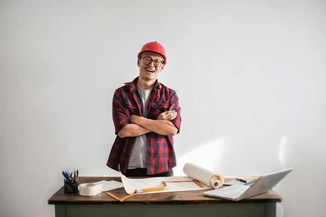 Smiling male contractor wearing a red hard hat and plaid shirt standing behind a desk with blueprints