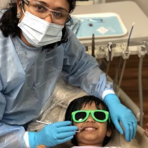 GILROY DENTAL SMILES Dentist examining a child's teeth. Both wear eye protection and masks. Bright green sunglasses on the child.