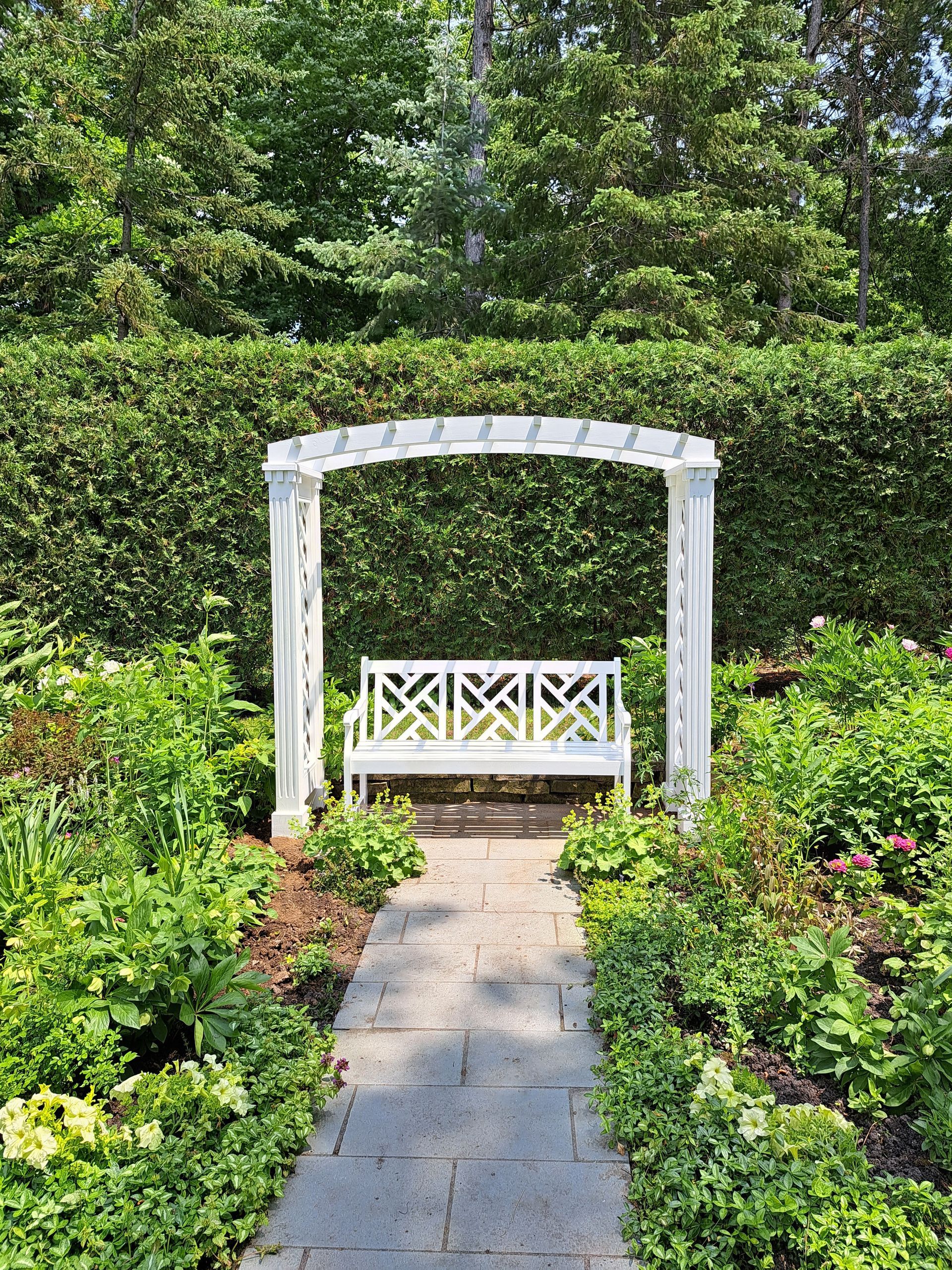 A white bench is sitting under a white arch in a garden.