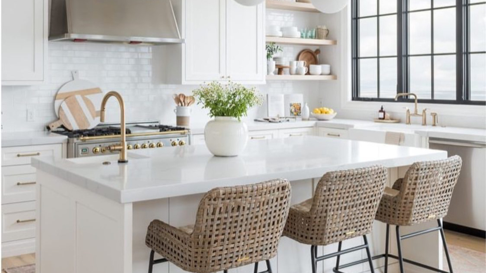 Bright white kitchen with marble island, woven bar stools, brass faucet, and open shelves.