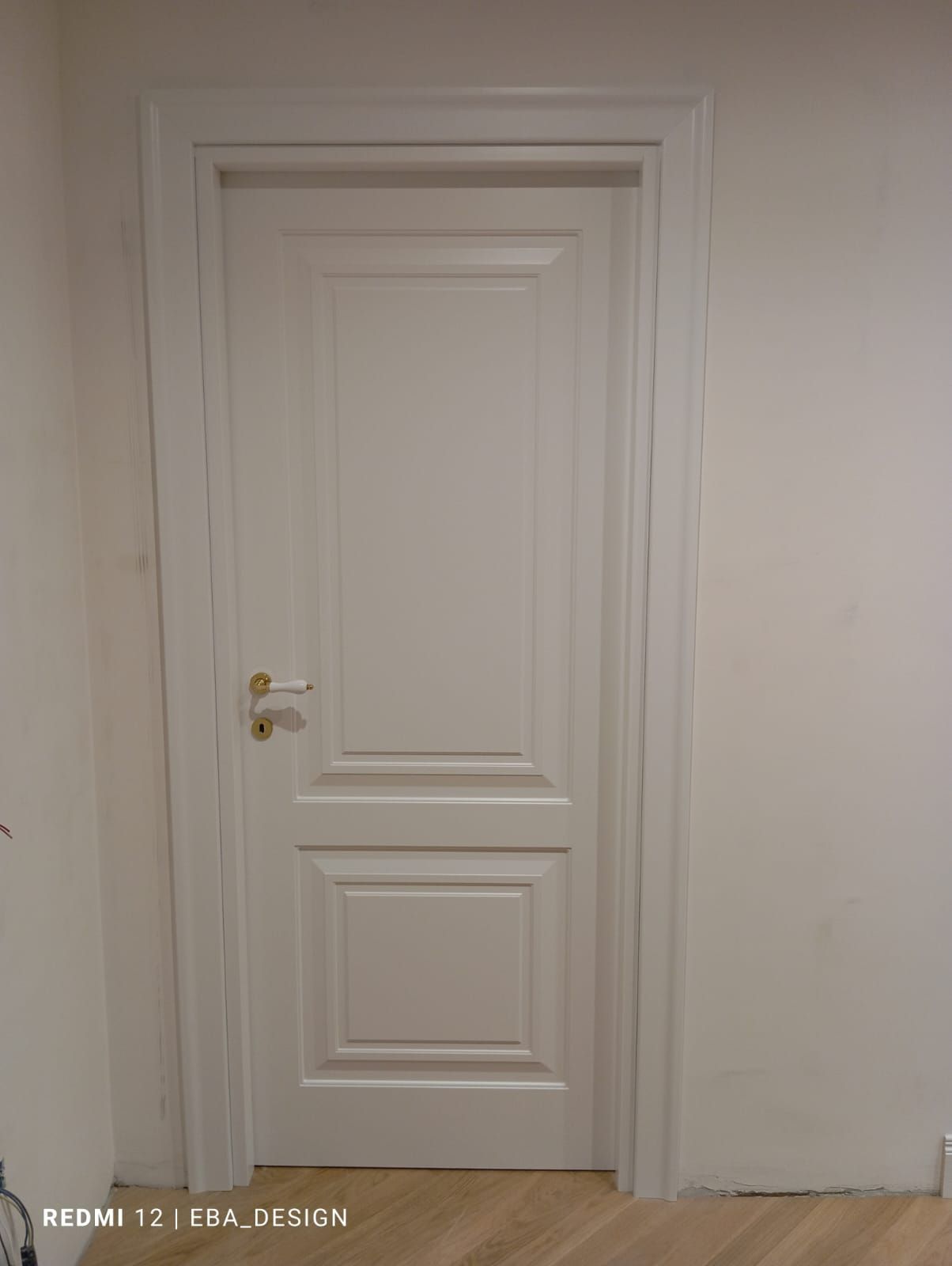 Porta interna con pannelli bianchi, maniglia e telaio dorati, contro una parete bianca.