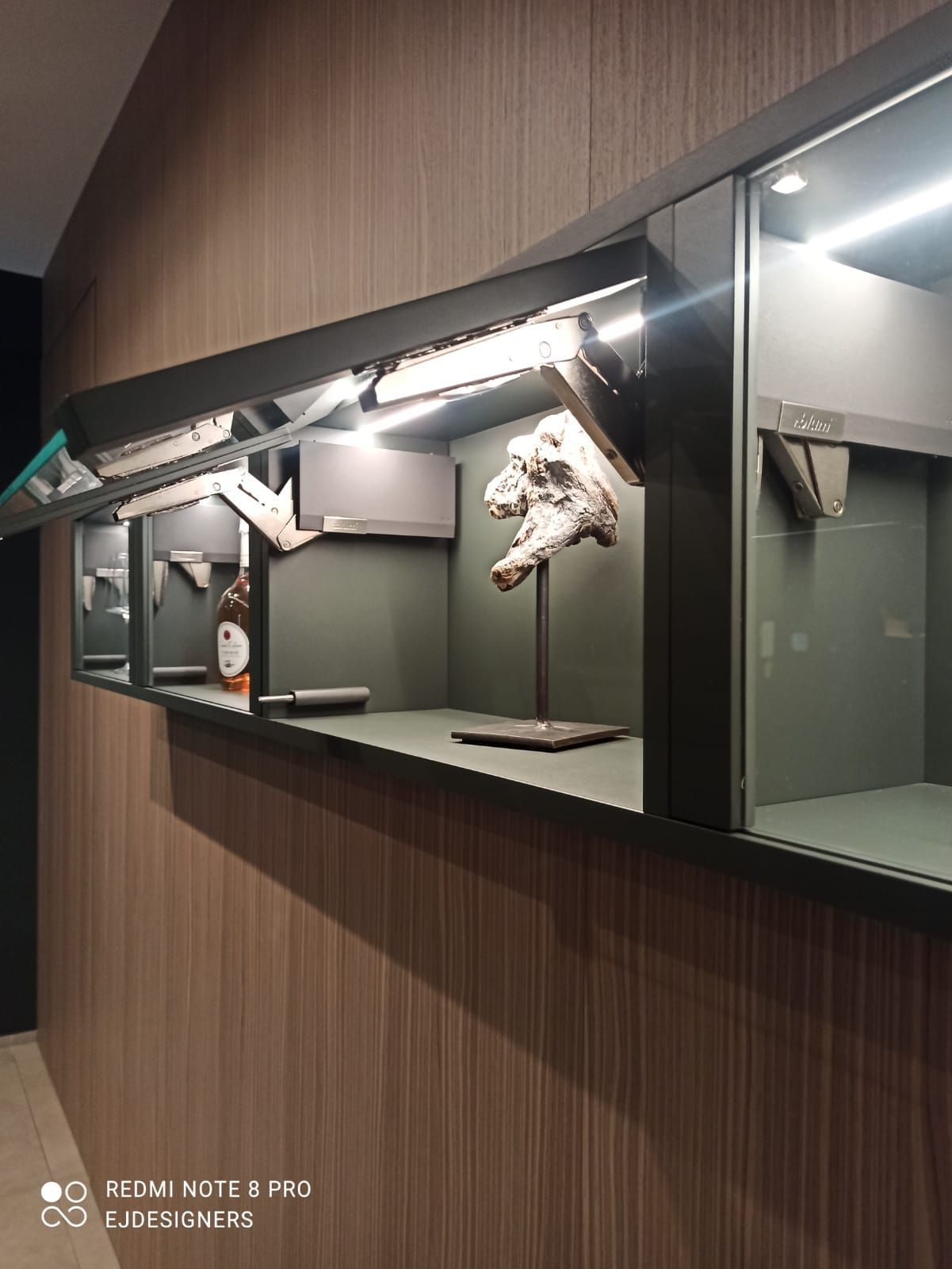 Vetrina a parete con elementi in legno. Presenta opere d'arte, illuminazione e interni scuri con pannelli in legno.