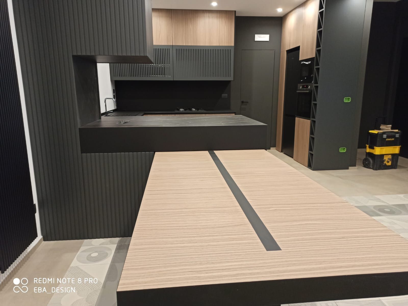 Cucina moderna con mobili e isola neri e in legno; è visibile un aspirapolvere.