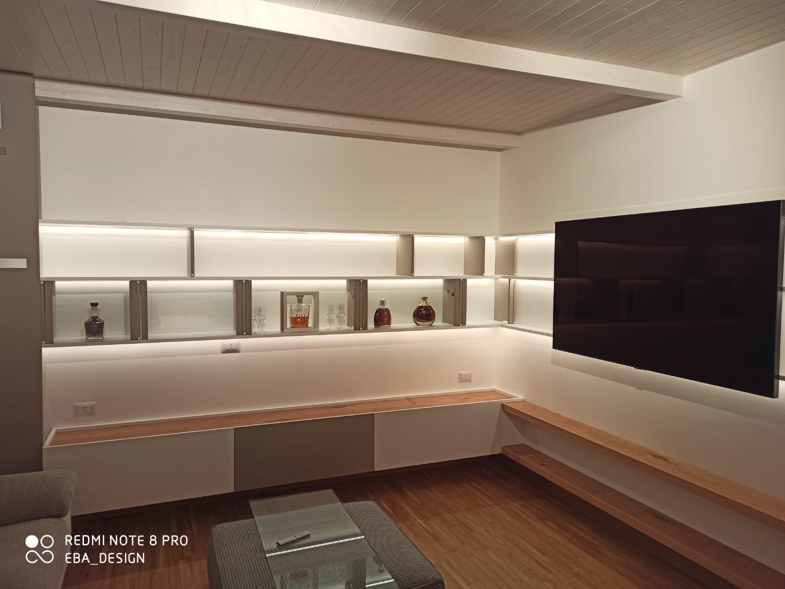 Soggiorno moderno con mensole integrate, TV e pavimento in legno. Pareti bianche, illuminazione a LED.