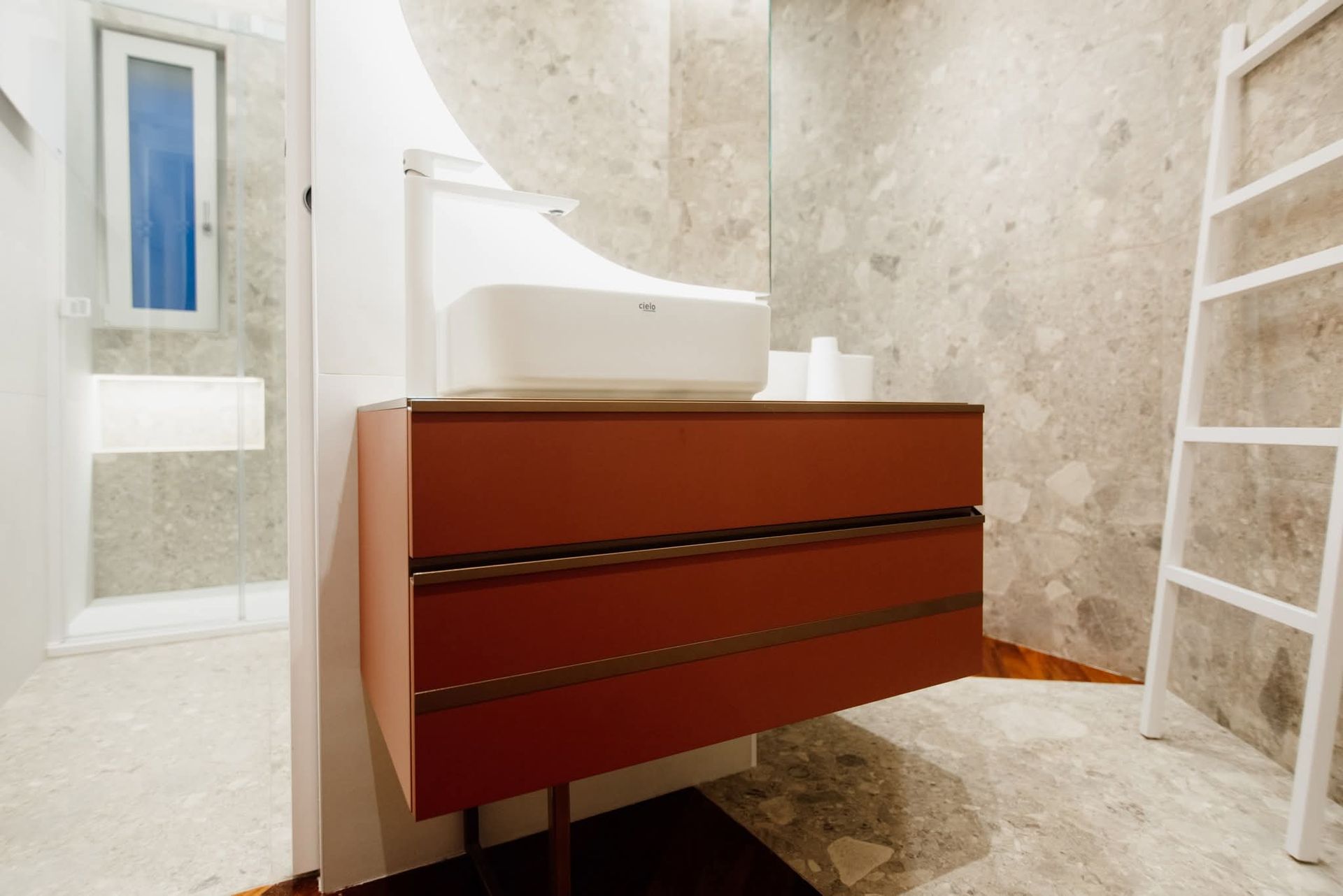 Bagno con mobiletto rosso, lavandino bianco e scala contro un muro di pietra.